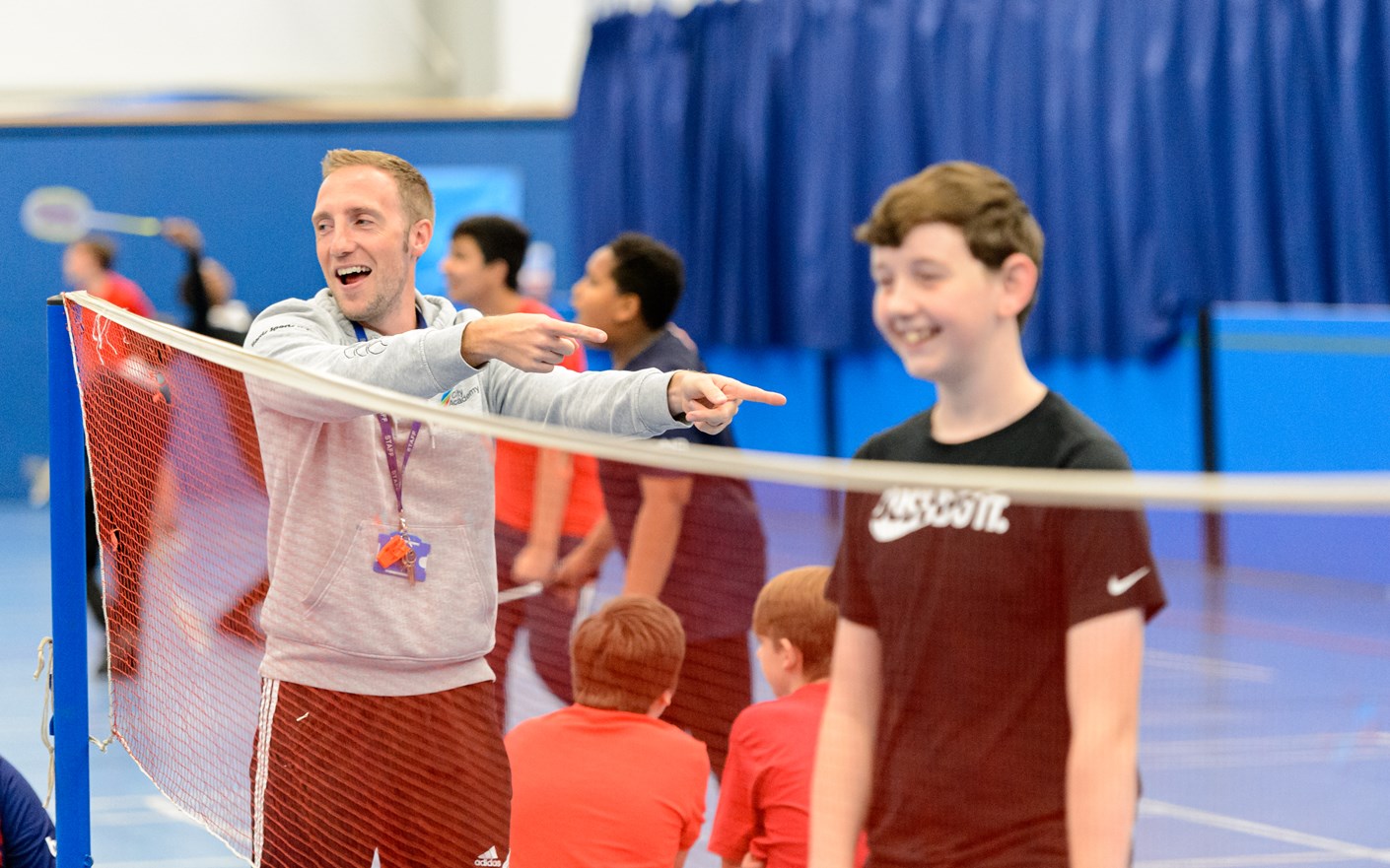 Badminton coach grinning and pointing at a smiling child player behind the net