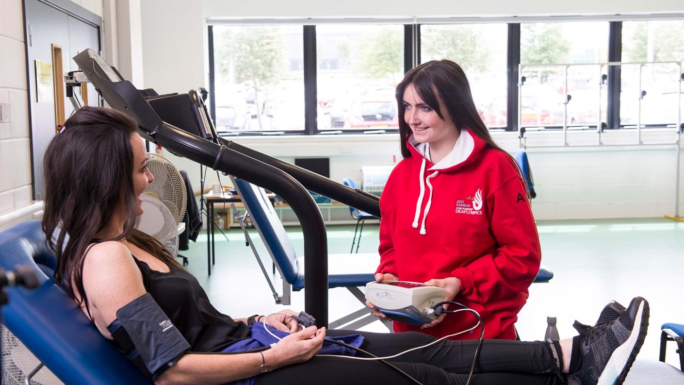 Dr Amanda Pitkethly is sat on an examination bed with a blood pressure monitor on her arm, which is monitored by a smiling student.